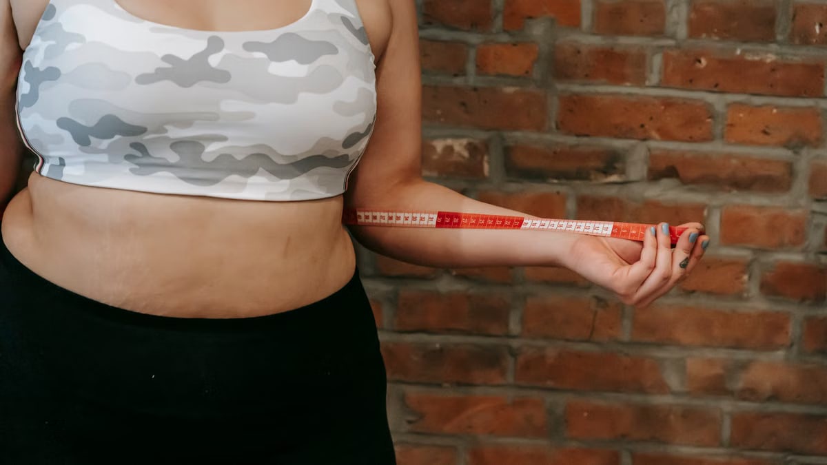 Crop anonymous plump female using measuring tape for measuring belly near brick wall