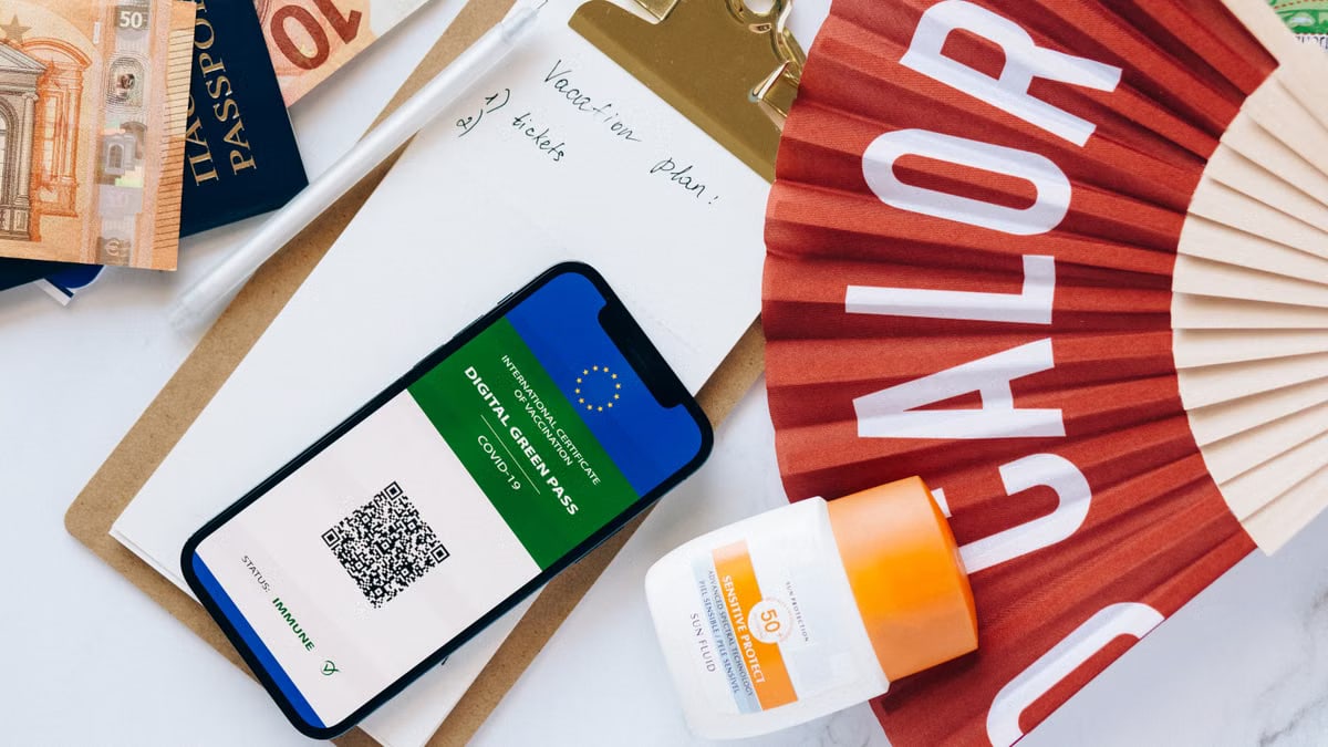 Flat lay of travel essentials featuring a digital green pass, passport, and other travel items on a table.